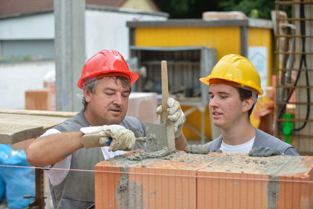 Zwei Bauarbeiter mit roten und gelben Schutzhelmen tragen frischen Mörtel auf gestapelte Ziegel auf einer Baustelle auf. Einer verwendet eine Kelle während der andere aufmerksam zuschaut.