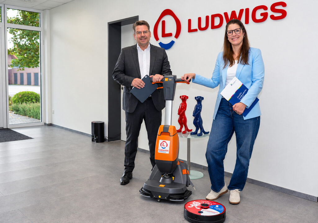 Eine Frau und ein Mann stehen in einem hellen Innenraum vor einer Wand mit dem Logo des Gebäudereinigungsunternehmens Ludwigs und lächeln in die Kamera. Zwischen ihnen befindet sich eine professionelle Reinigungsmaschine für das Gebäudereiniger Handwerk und davor ein runder Reinigungsroboter.